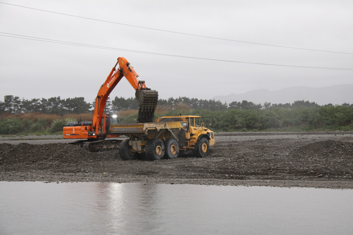 A digger dropping gravel into the bed of a truck on the bank of a river